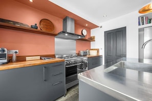 A modern kitchen with grey cabinetry, a stainless steel island, and a built-in oven. - Knutsford Home | Sun Trap Garden | Sleeps 8 (Cheshire East)