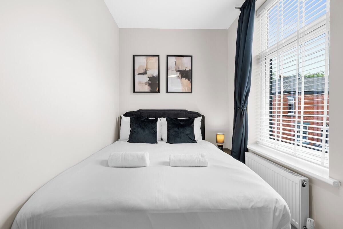 Une chambre d’hôtel avec un lit, deux œuvres d’art encadrées au mur, une fenêtre avec des stores et une lampe de chevet.