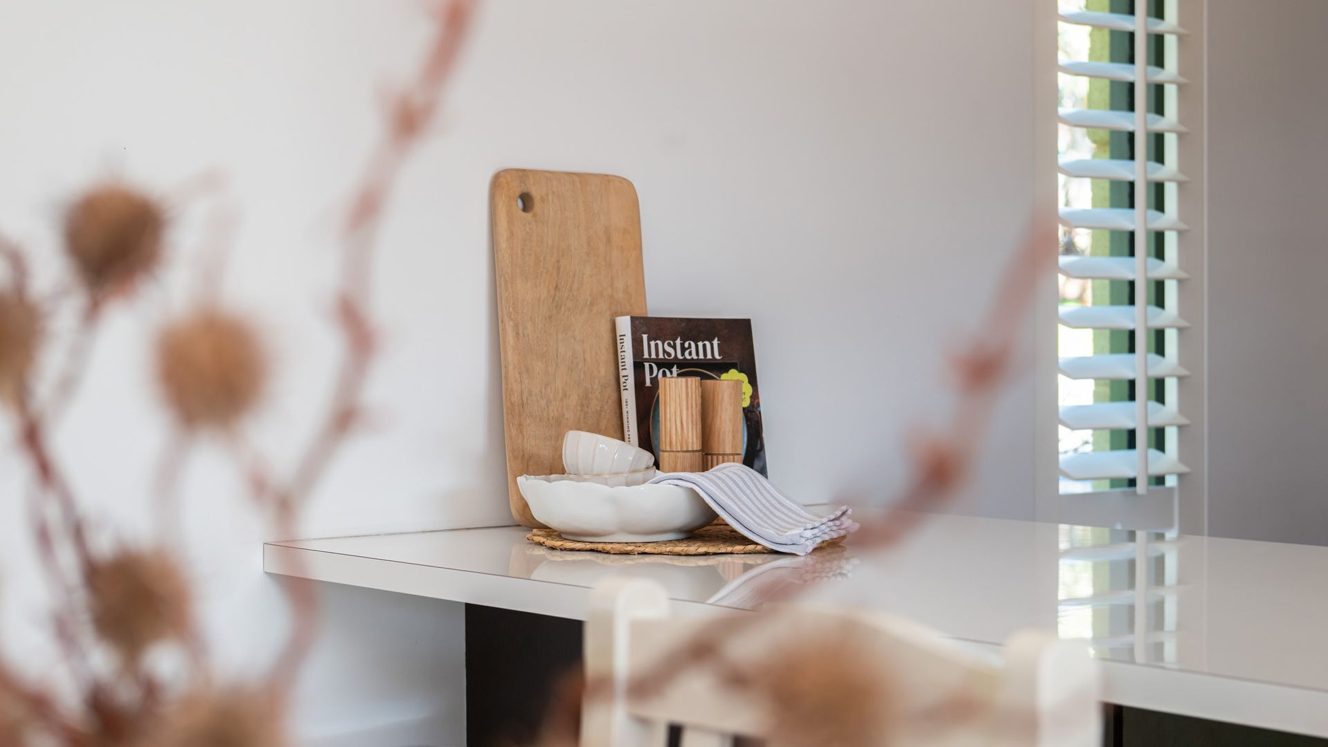 Uma bancada de cozinha com uma tábua de corte de madeira, um livro de receitas, uma tigela branca e um pano listrado.