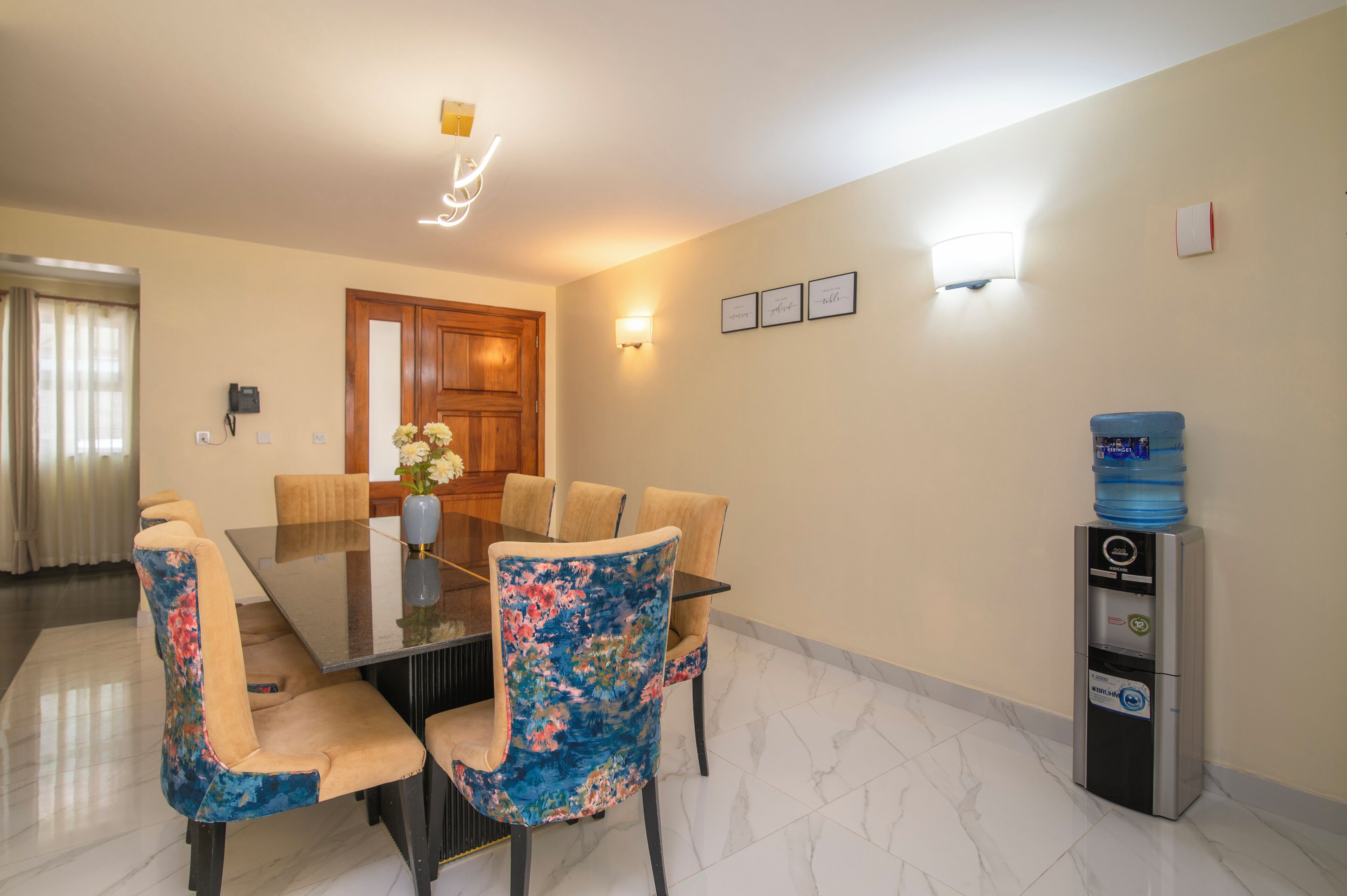 A dining area with a glass table, floral chairs, a water dispenser, and framed wall art.