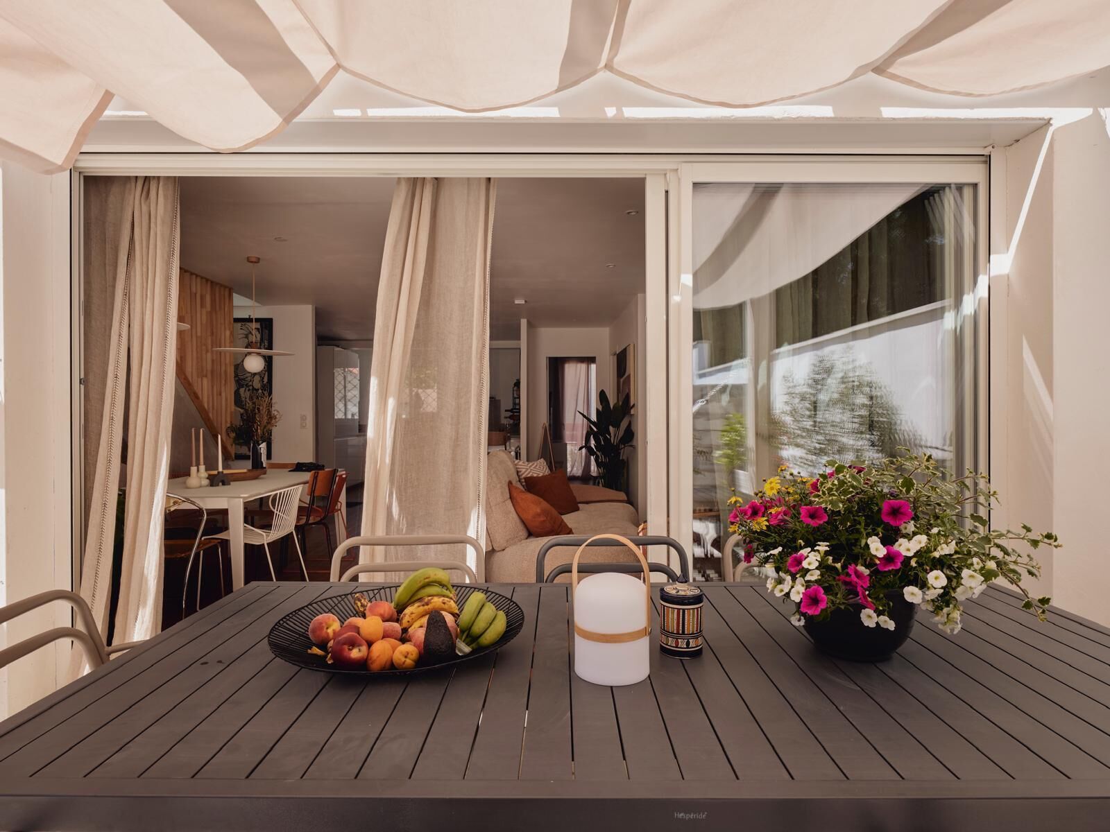 A dining table with a fruit bowl, a candle, and a flower vase in a modern outdoor setting.