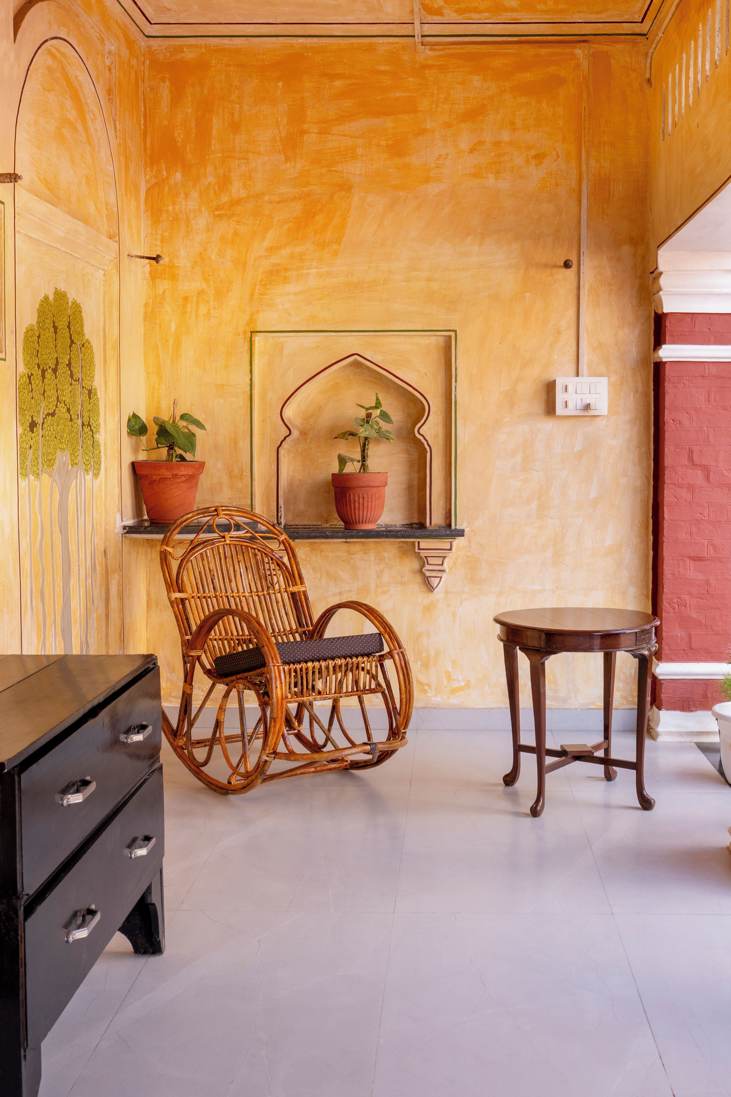 A room with a wicker chair, two potted plants, a small table, and a black dresser.