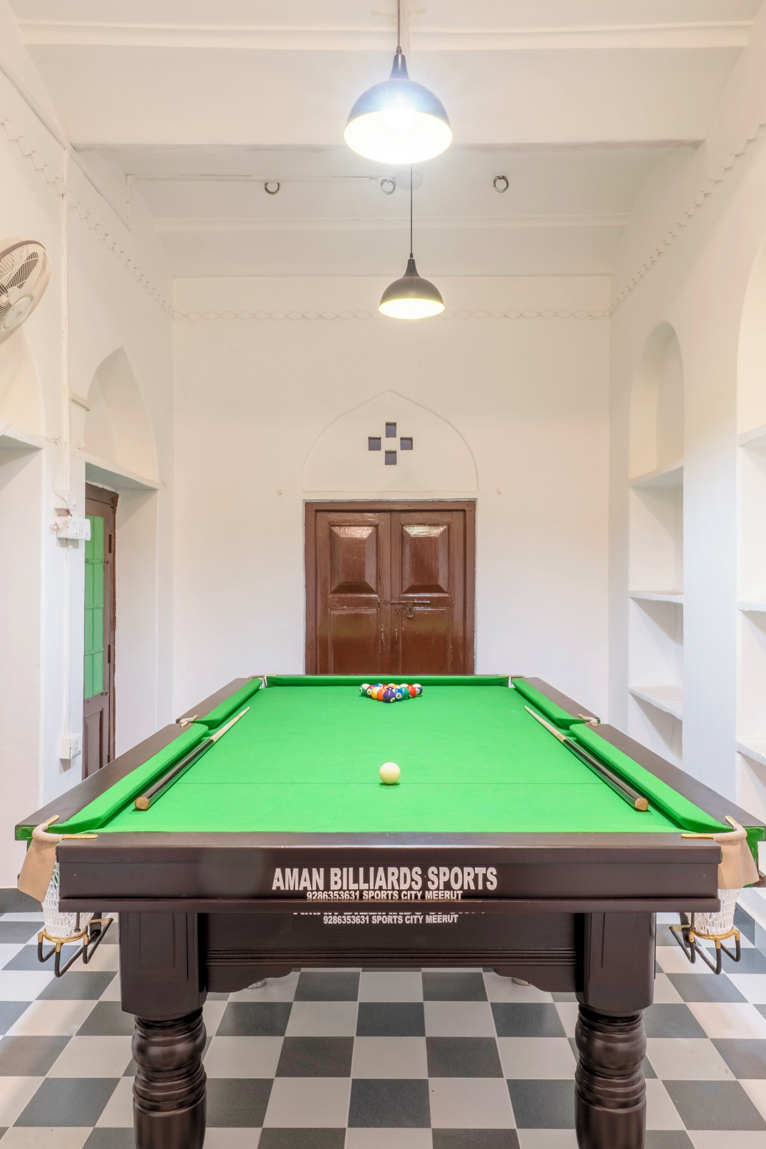 A pool table with a green felt surface, a white ceiling with a hanging light fixture, and a wooden door with a small window.