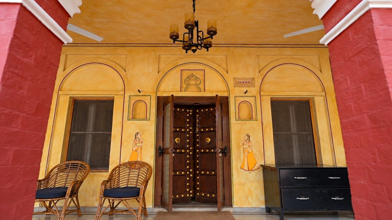 A traditional building entrance with a wooden door, two wicker chairs, and a black cabinet.
