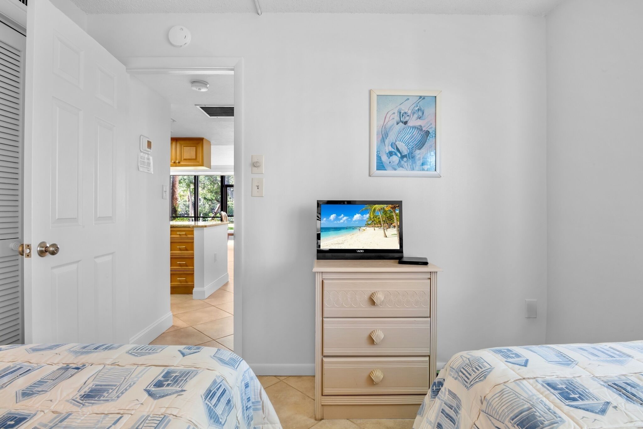 A bedroom with two beds, a TV on a dresser, and a view of a beach through a window.