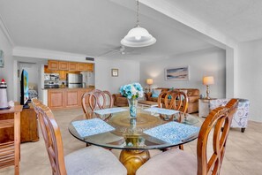 A dining area with a glass table surrounded by wooden chairs, a vase with flowers, and a kitchen in the background. - Midnight Cove II – Unit 513 - Pool View! (Siesta Key)