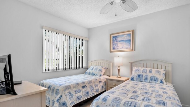 A bedroom with two beds, a desk, a chair, a ceiling fan, a window with blinds, and a painting on the wall. - Midnight Cove II – Unit 513 - Pool View! (Siesta Key)