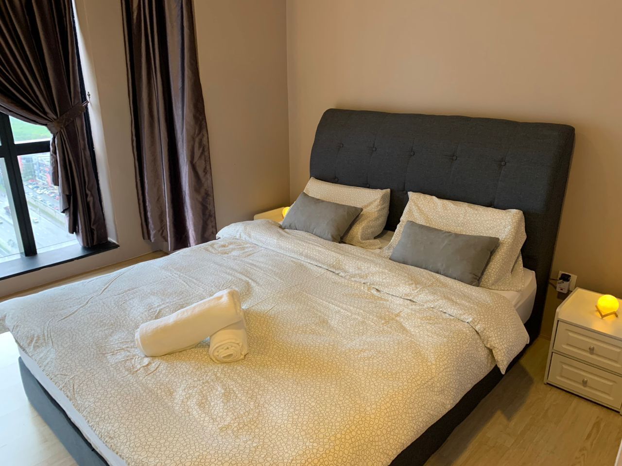 Een net opgemaakt bed met een grijs hoofdeinde, een wit kussen en een opgerolde witte handdoek op het bed.