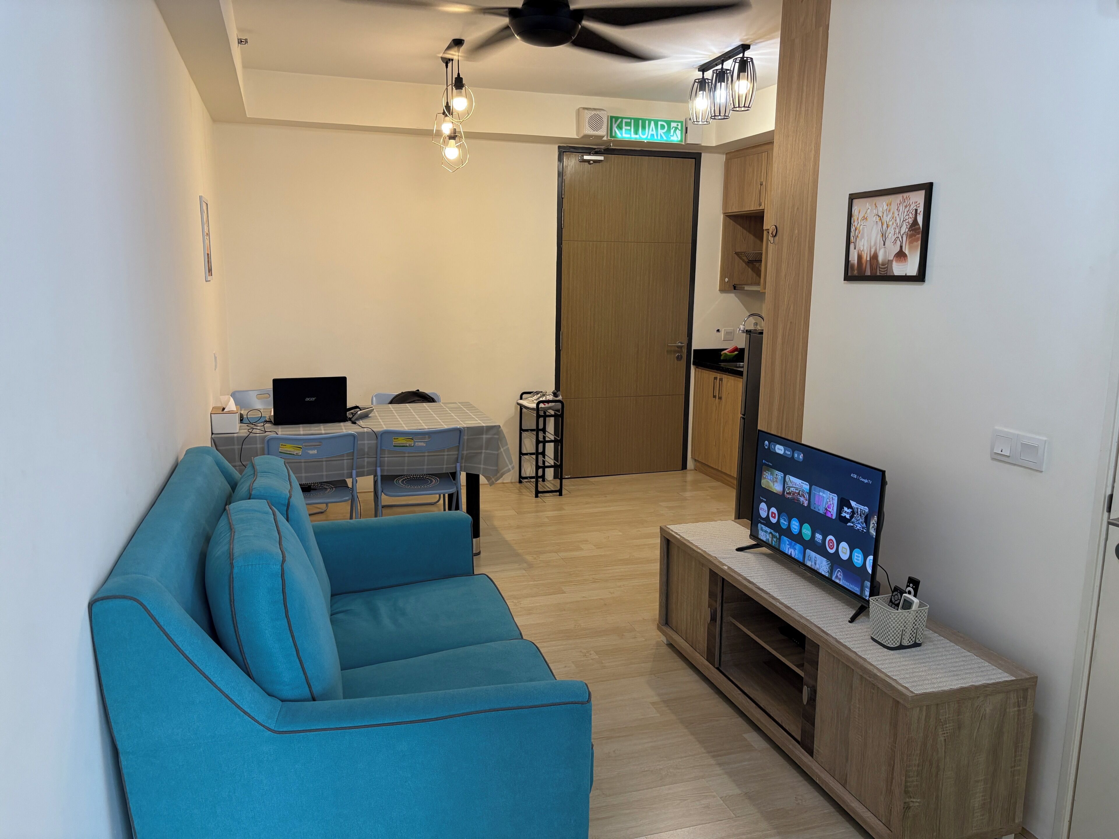 Una sala de estar con un sofá azul, un televisor sobre un mueble de madera, un comedor con una mesa y sillas, y una cocina con armarios y un refrigerador.