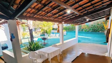 Un espacio al aire libre techado con una piscina, sillas de plástico blancas y una mesa.