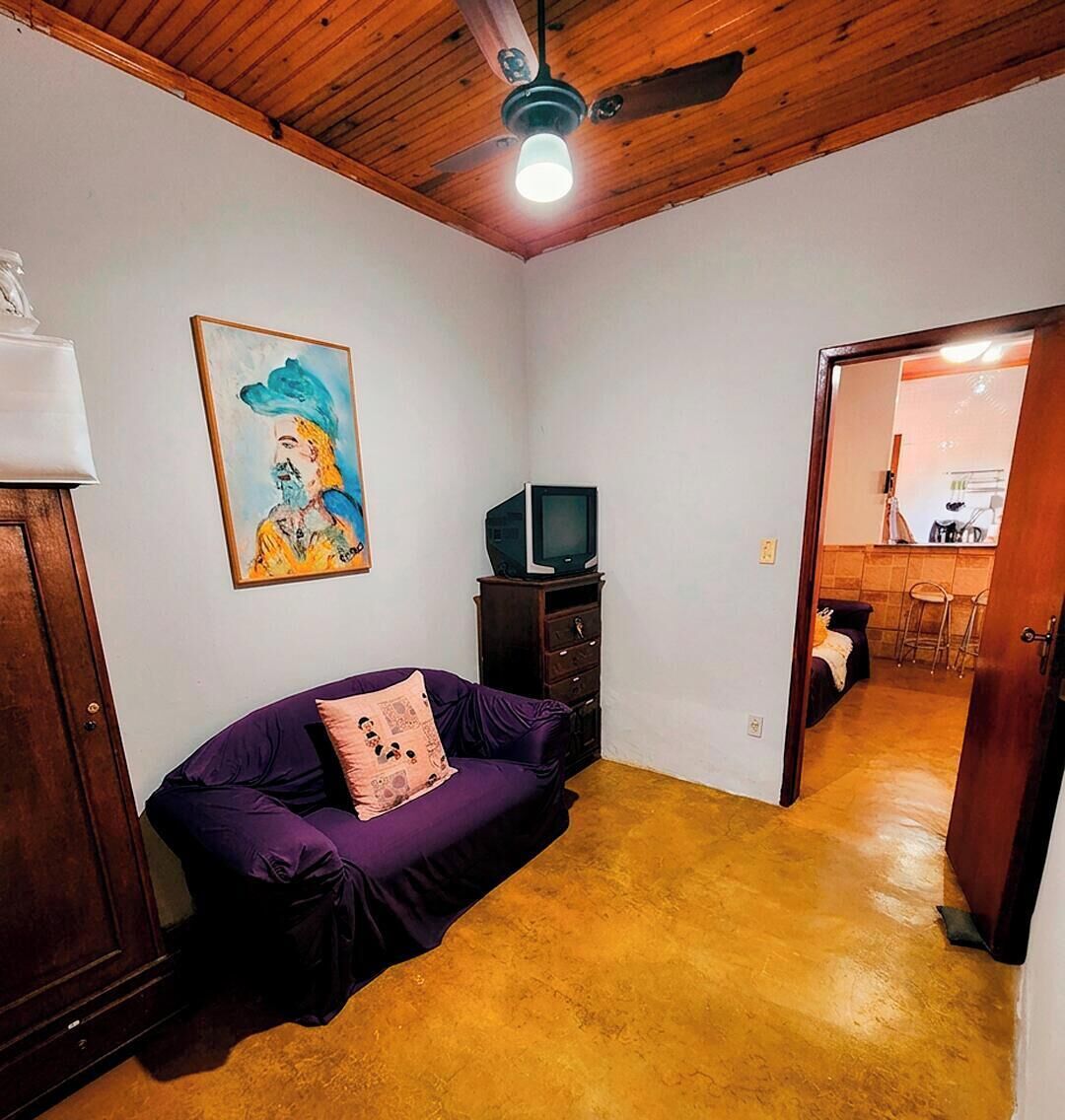 Una habitación con un sofá púrpura, un televisor sobre un mueble de madera, un ventilador de techo y un cuadro en la pared.