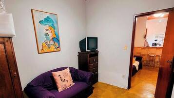 Una habitación con un sofá púrpura, un televisor sobre un mueble de madera, un ventilador de techo y un cuadro en la pared.