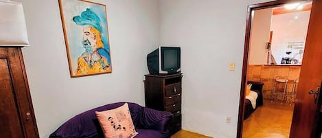 Una habitación con un sofá púrpura, un televisor sobre un mueble de madera, un ventilador de techo y un cuadro en la pared.