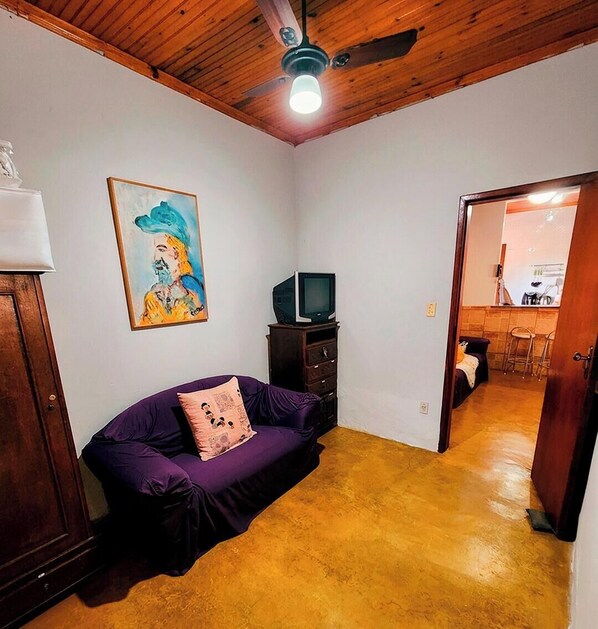 A room with a purple sofa, a television on a wooden cabinet, a ceiling fan, and a painting on the wall.