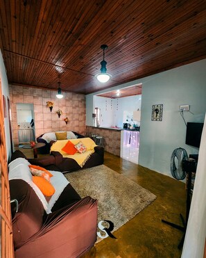 A cozy living room with a sofa, a coffee table, and a rug. There is a kitchen in the background with a counter and a fan.
