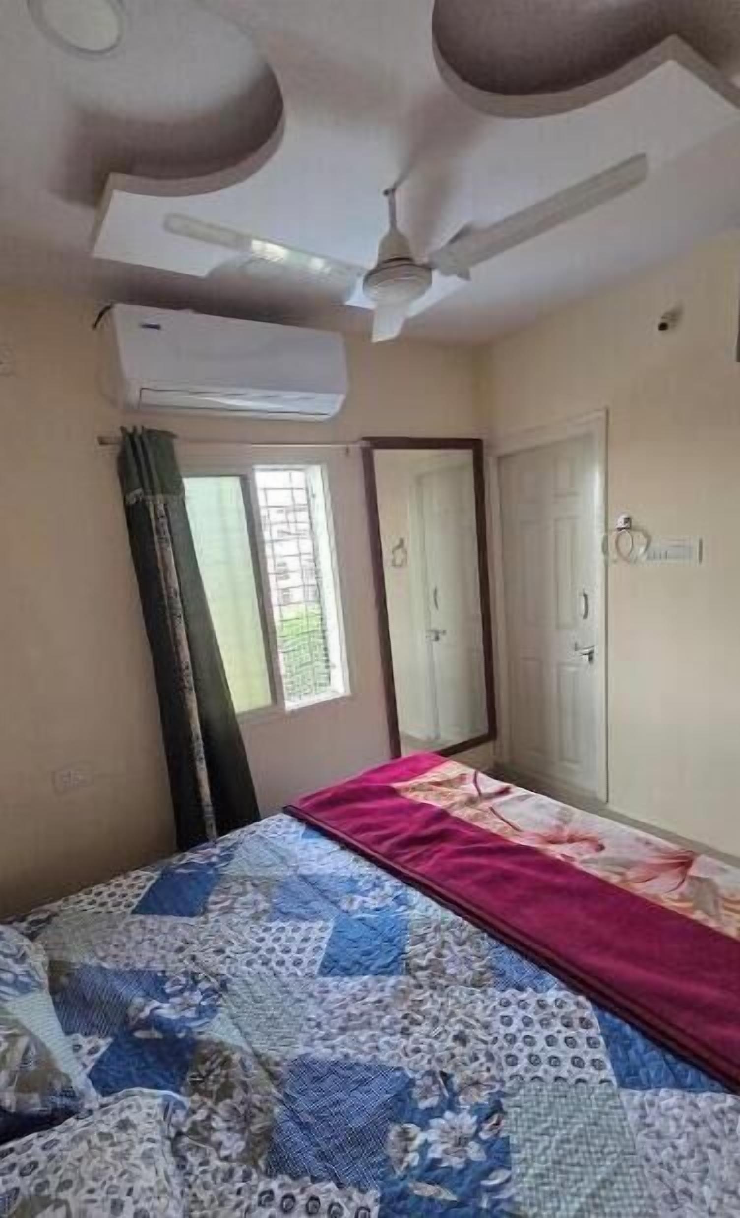 A bedroom with a bed, an air conditioning unit, a ceiling fan, a window with curtains, and a door.