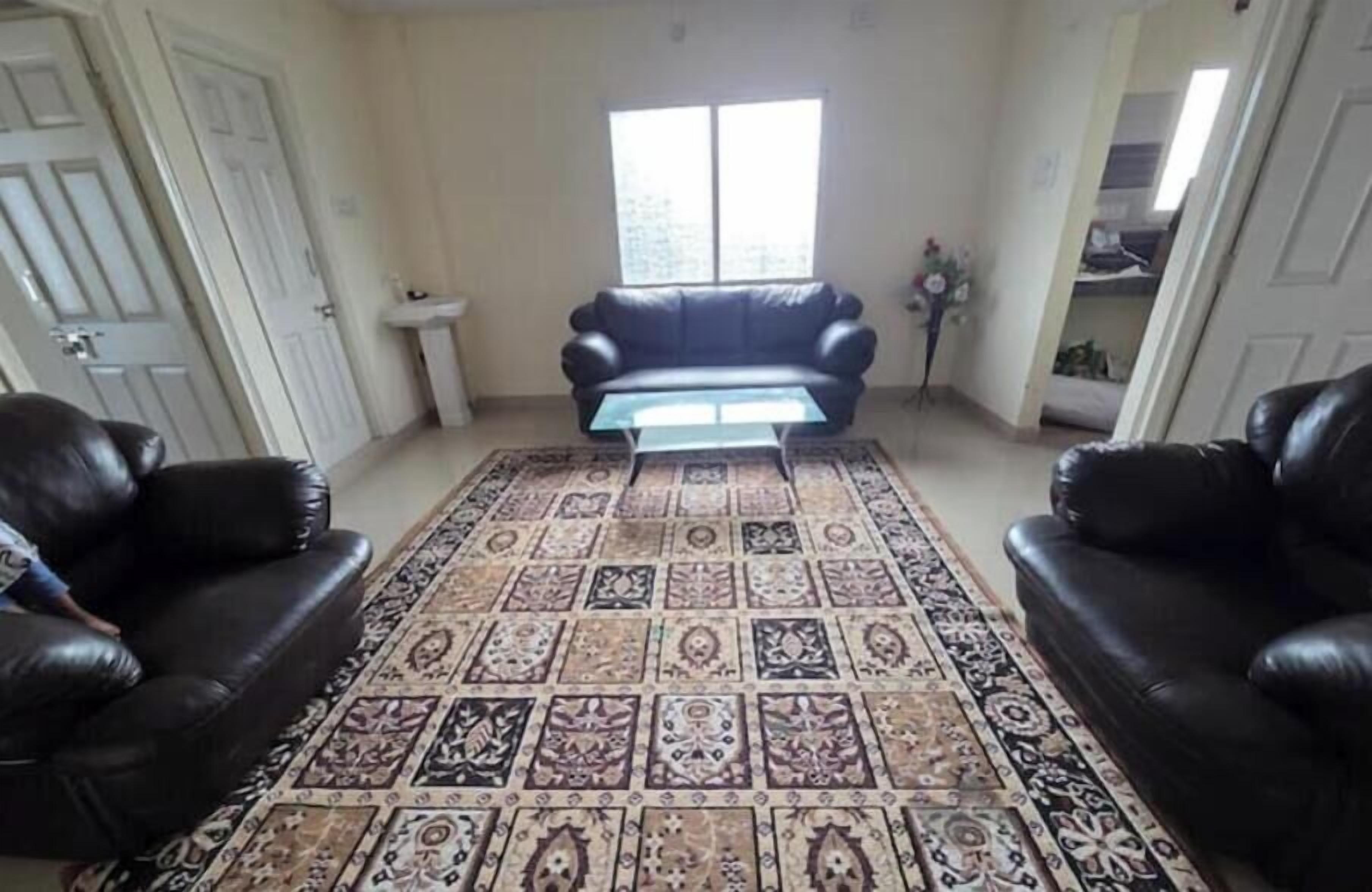 Un salon avec un tapis à motifs, deux canapés en cuir noir, une table basse et une fenêtre avec des stores.