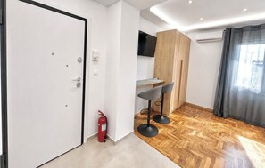 Une pièce moderne avec un plancher en bois, une porte blanche, un extincteur, un petit coin bar avec des tabourets et un téléviseur fixé au mur.