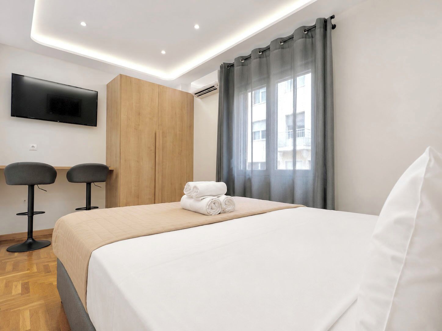 Habitación de hotel con cama, televisor montado en la pared, dos taburetes de bar y una ventana con cortinas.