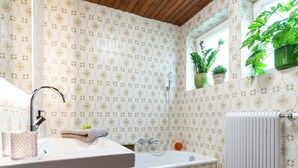 A bathroom with a wooden ceiling, patterned wallpaper, a sink with a mirror, a shower, and a radiator.