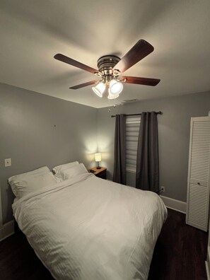 Un dormitorio con cama, mesita de noche, ventilador de techo y ventana con cortinas.