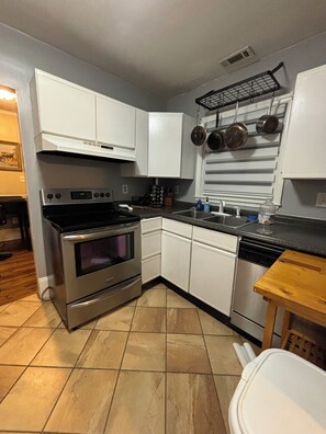 A kitchen with white cabinets, a stainless steel oven, a dishwasher, and a window with hanging pots. - Charming 2BR Bungalow/Walkable Chantilly & Plaza Midwood Front Half of Home (Charlotte)
