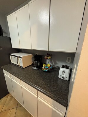 Una kitchenette con armarios blancos, un microondas, una cafetera, un tostador y una cesta de aperitivos.