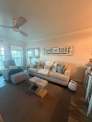 Een woonkamer met een bank, fauteuil en poef, een plafondventilator, en een bord aan de muur met de tekst 'SAND AND SURF'.