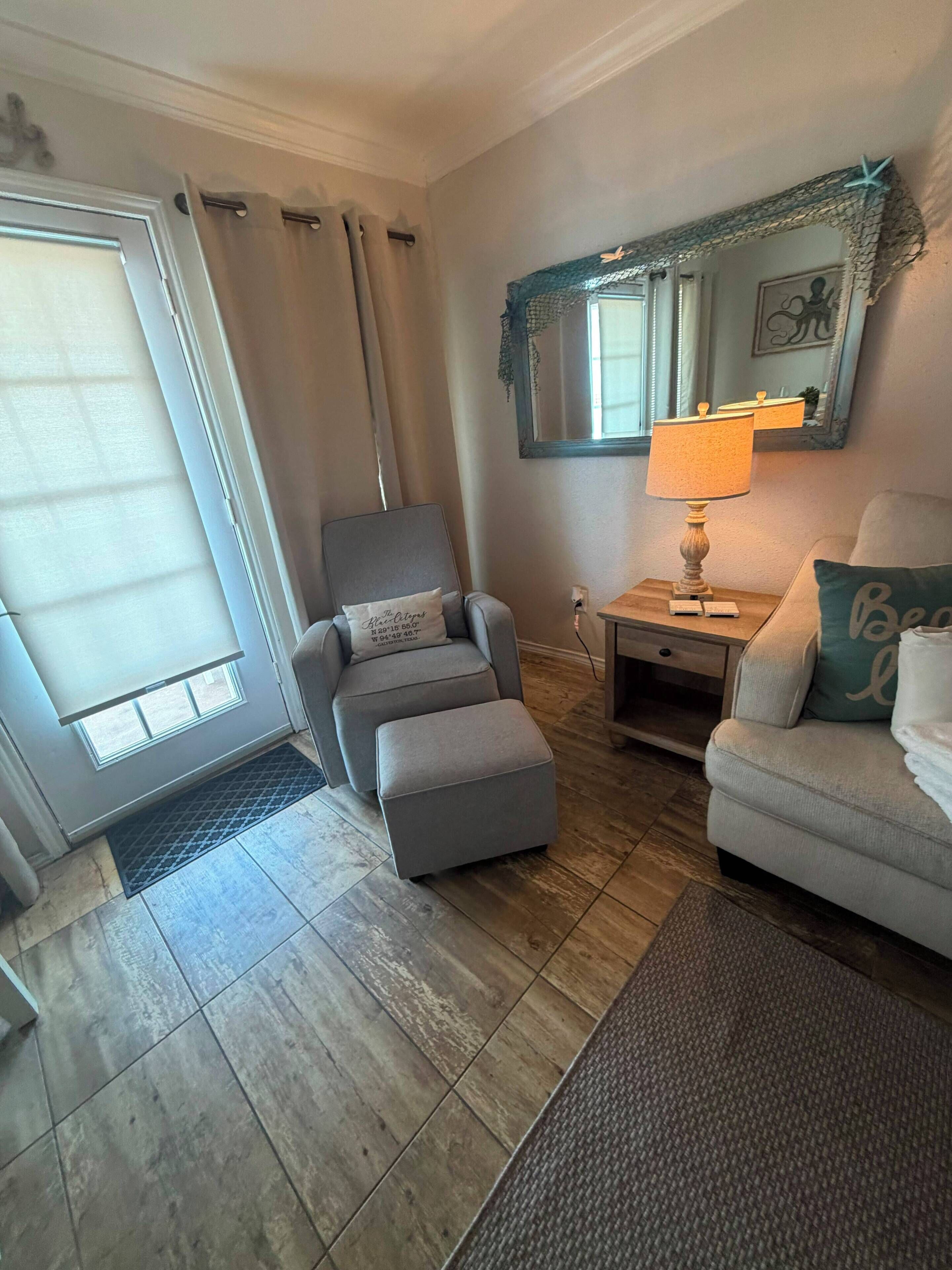 A living room with a grey armchair, ottoman, a side table with a lamp, a sofa with a pillow, a large mirror, and a sliding glass door.