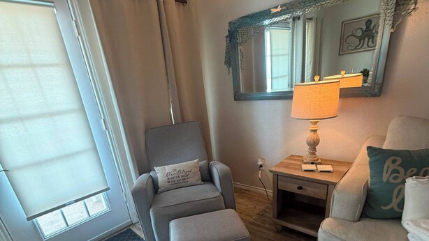 A living room with a grey armchair, ottoman, a side table with a lamp, a sofa with a pillow, a large mirror, and a sliding glass door. - Ocean Front Condo - 2 pools hot tub Cruise Parking on seawall babes beach (Galveston)