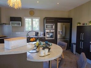 A modern kitchen with a built-in oven, microwave, and refrigerator. There is a round table with chairs and a bread box. - The Villa Under the Stars – Les Issambres (Roquebrune-sur-Argens)