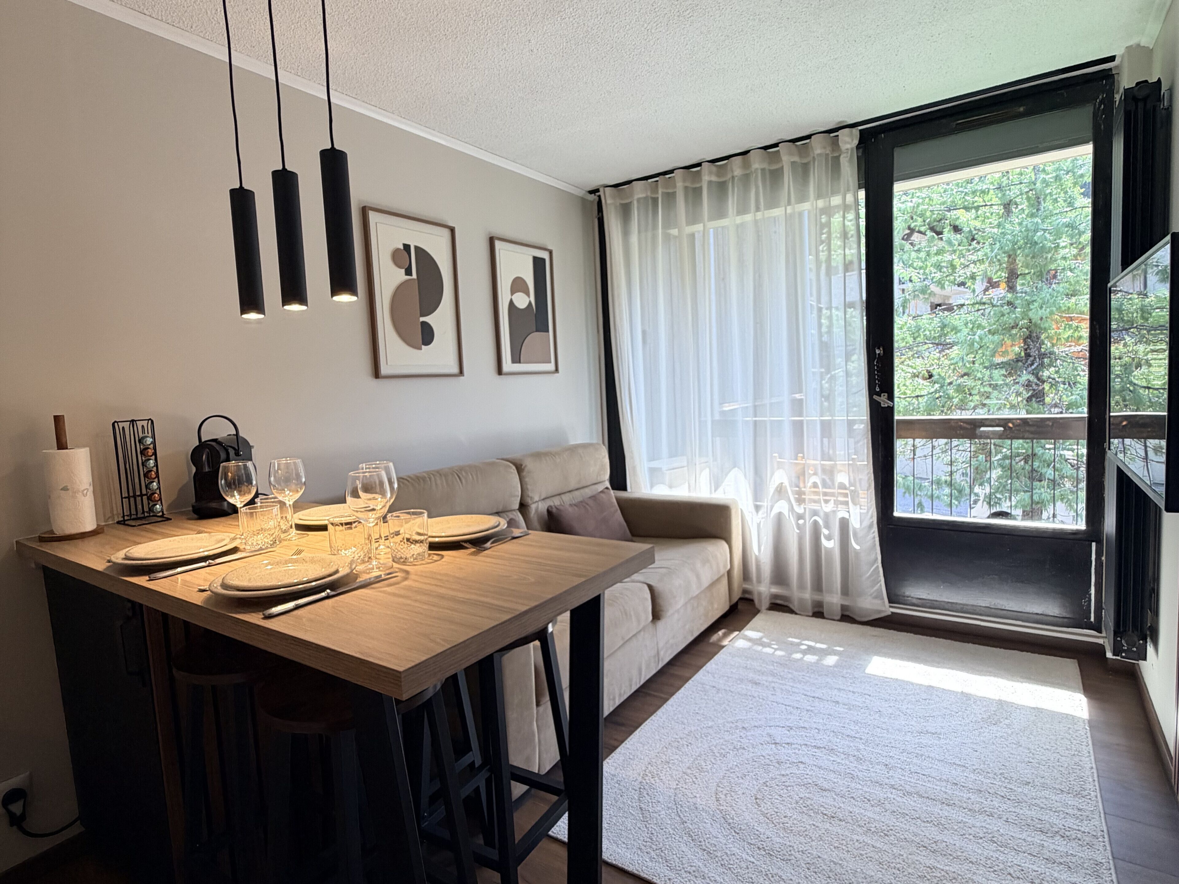 A dining area with a wooden table set for four, a sofa, and a large window with a view of greenery.