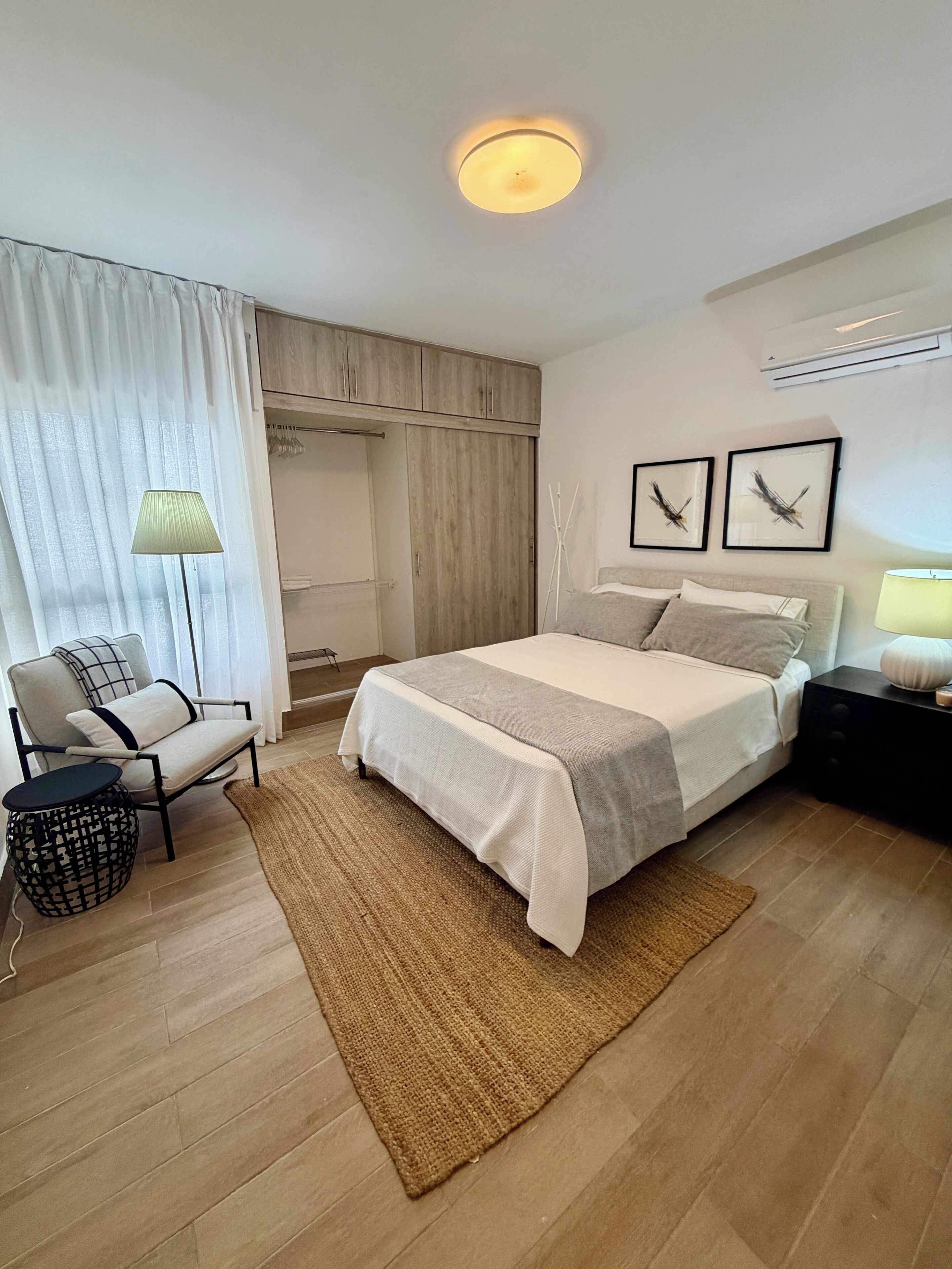 Habitación de hotel con cama, sillón, mesita de noche, lámpara, armario y dos cuadros en la pared.