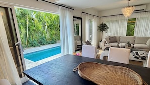 A modern living room with a pool view, a sofa, a coffee table, and a dining area with a black table and a woven placemat.