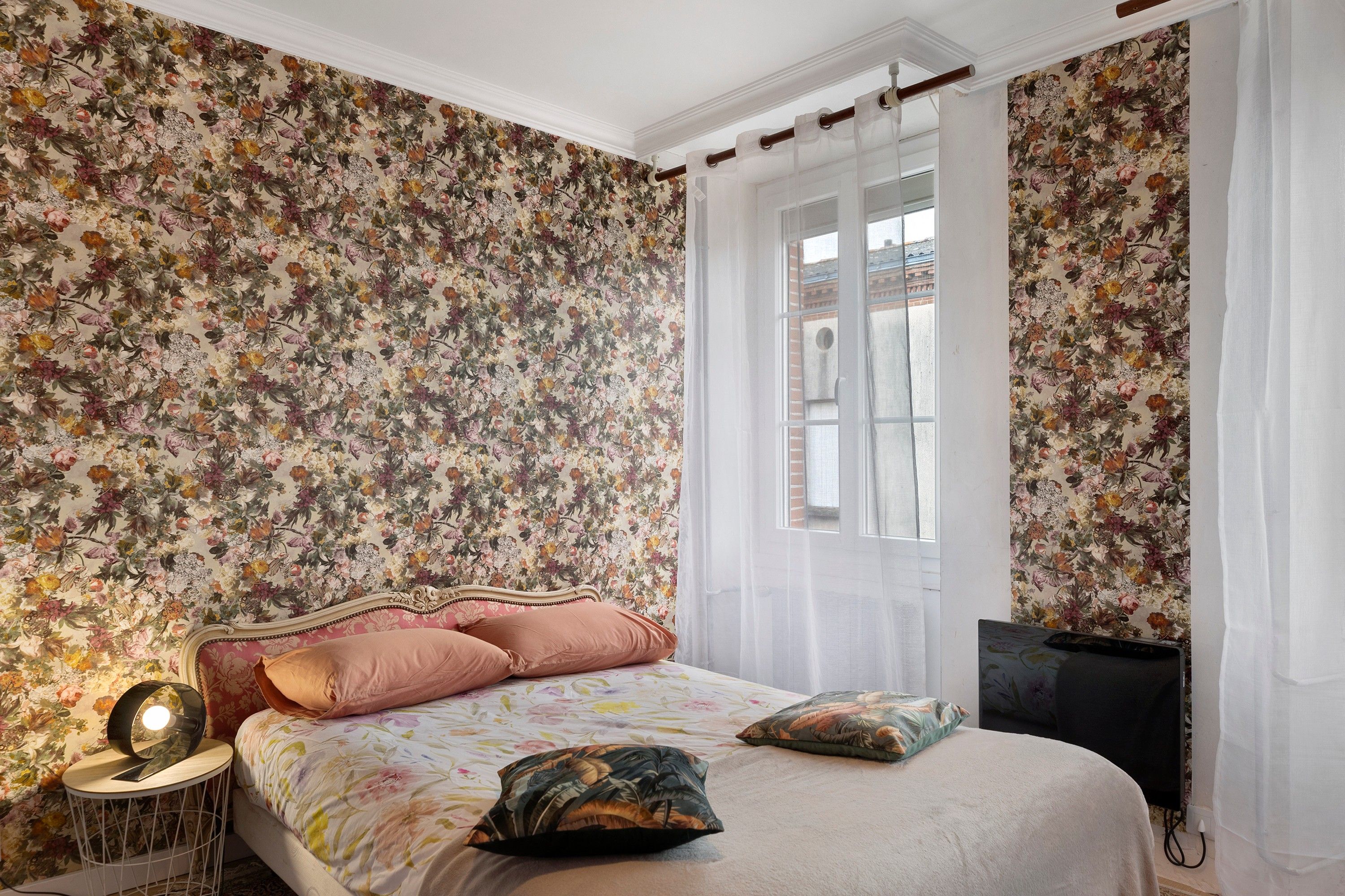 Une chambre avec un papier peint à motifs floraux, un lit avec de nombreux coussins, une table ronde et un téléviseur.