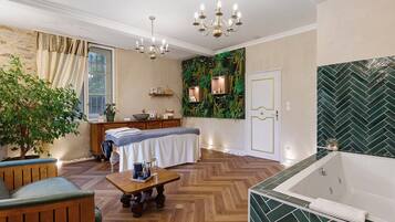 A well-lit room with a wooden floor, a large window with curtains, a chandelier, a massage table, a wooden bench, a small table with items, and a green wall with a herringbone pattern.