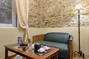 A room with a stone wall, a green leather sofa, a wooden table with a teapot and cups, and a floor lamp.