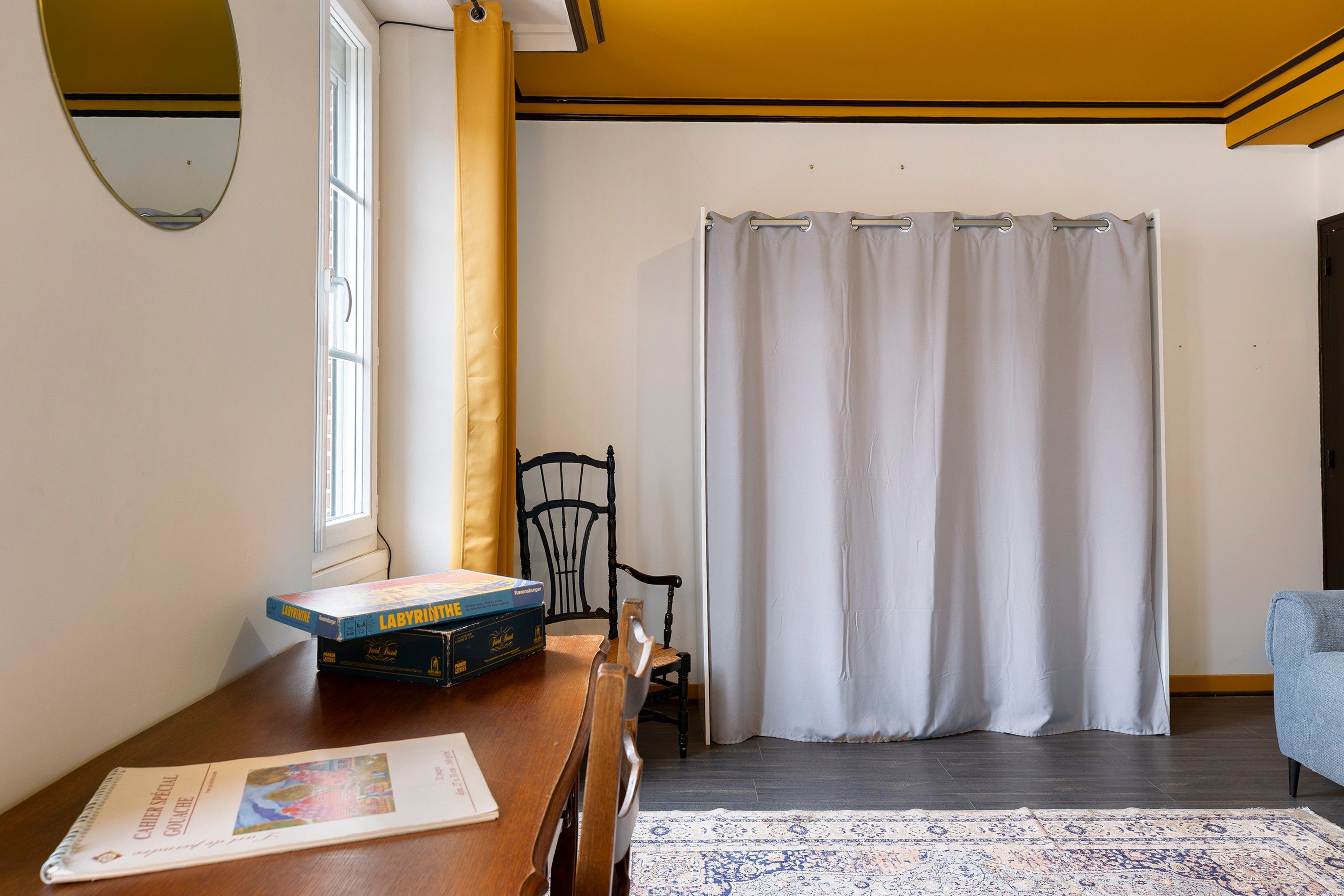 Ein Zimmer mit einem Holztisch, einem Stuhl, einem Fenster mit Vorhängen und einem gemusterten Teppich.