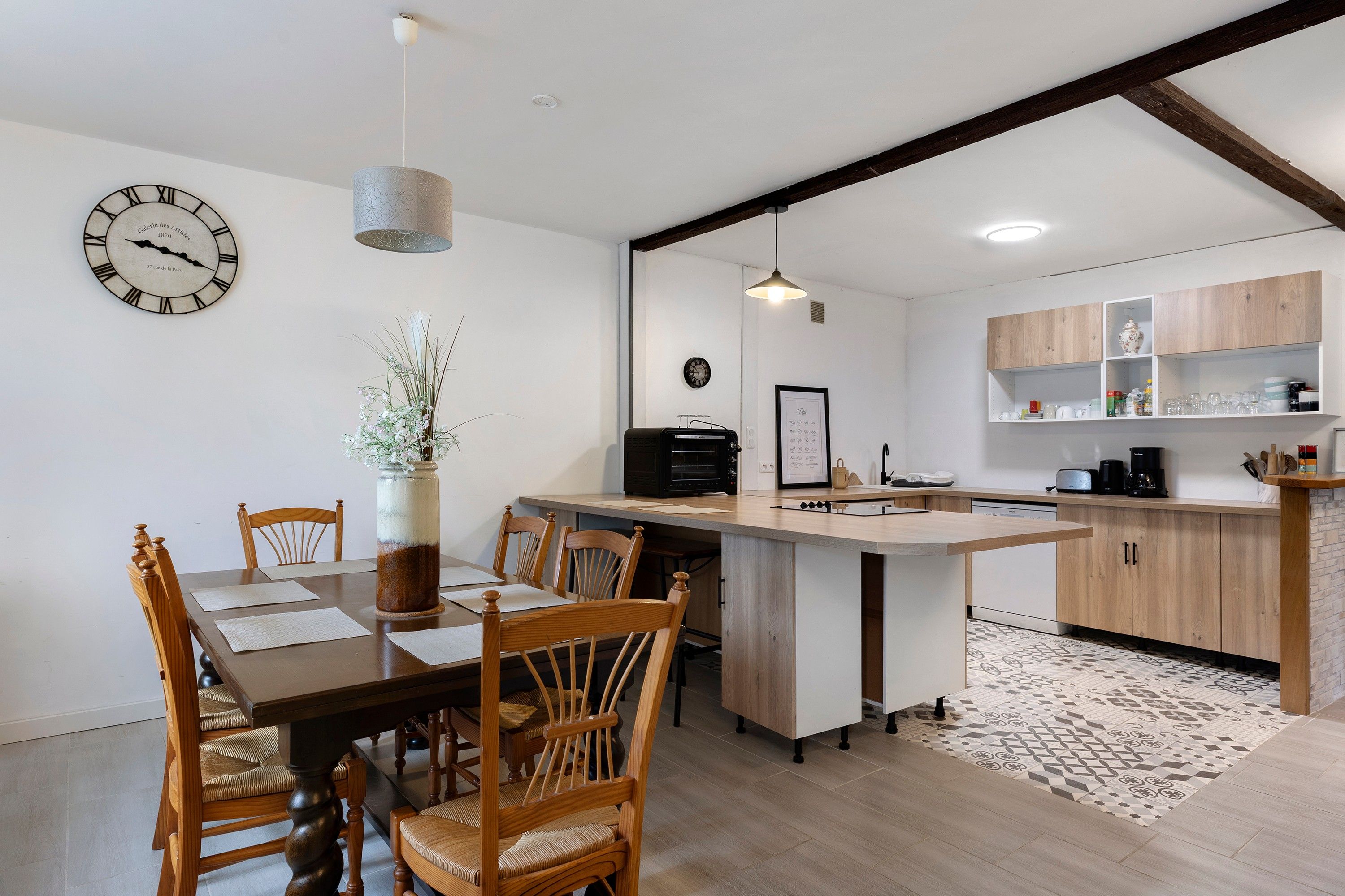 A modern kitchen with wooden cabinets, a dining table with chairs, a clock, and a hanging light fixture.