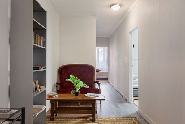 A narrow hallway with a bookshelf, a red armchair, a wooden table with a plant, and a bed in the background.