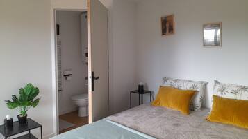 Une chambre à coucher comprenant un lit, une table de chevet, une plante en pot, des toilettes et une porte donnant sur une salle de bain.