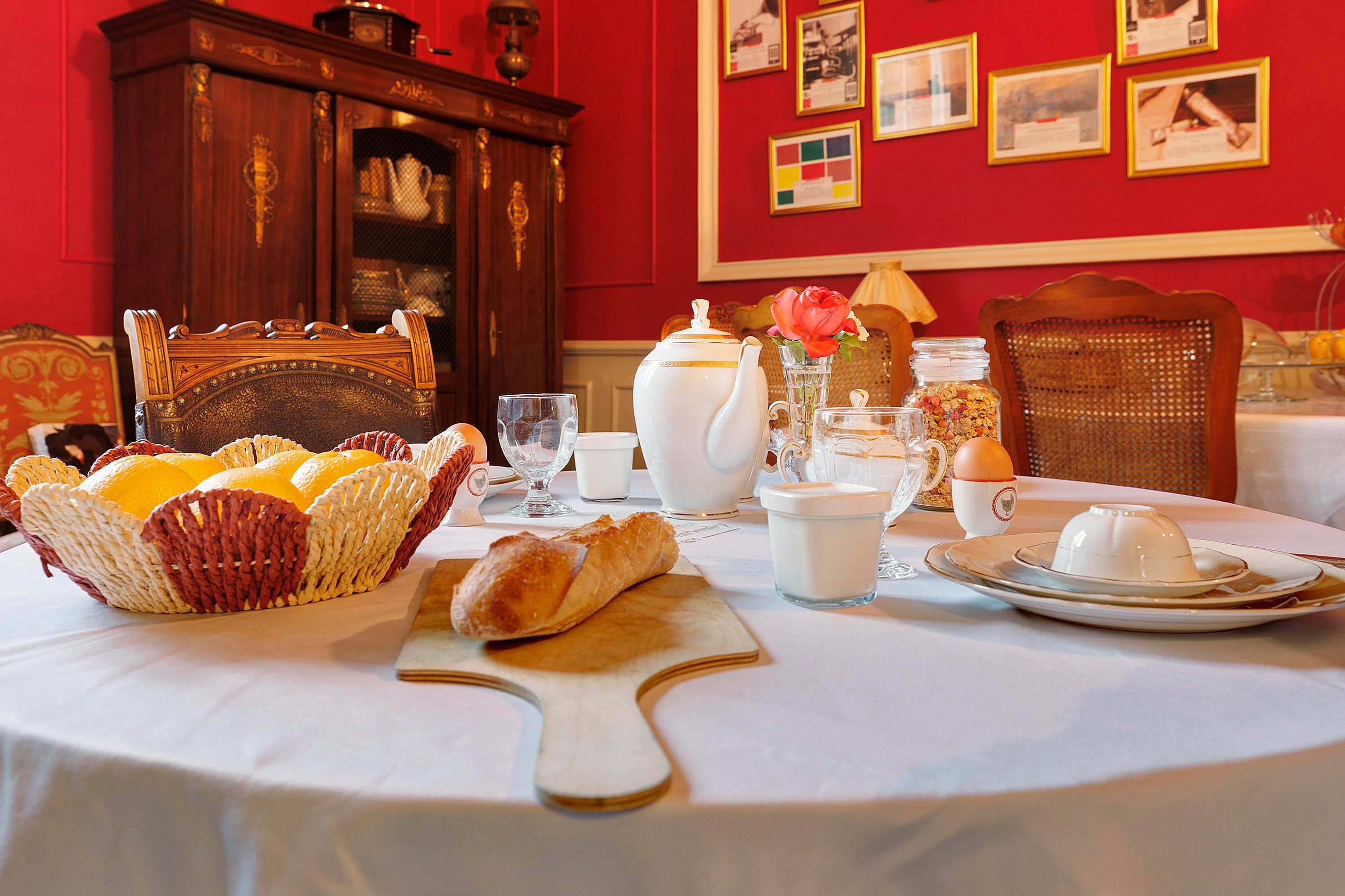 Un desayuno con una mesa dispuesta para dos, que incluye pan, pasteles, una tetera, tazas y un jarrón con flores.
