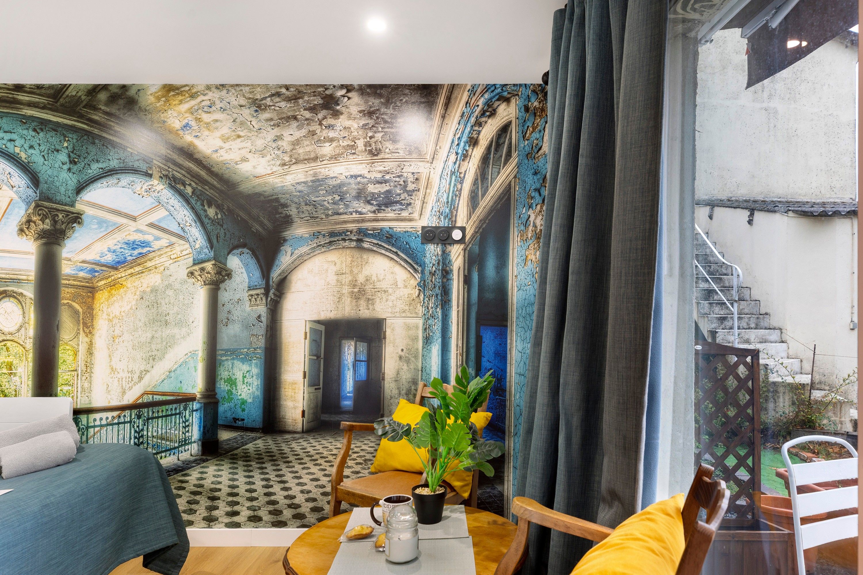 Una habitación con un mural de un edificio antiguo, una mesa de comedor con sillas, una planta y una escalera visible a través de una ventana.
