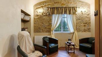 A cozy room with stone walls, a window with curtains, two leather chairs, a small table with a bottle, and a radiator.