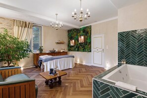 A well-lit room with a wooden floor, a large window with curtains, a chandelier, a massage table, a wooden bench, a small table with items, and a green wall with a herringbone pattern.