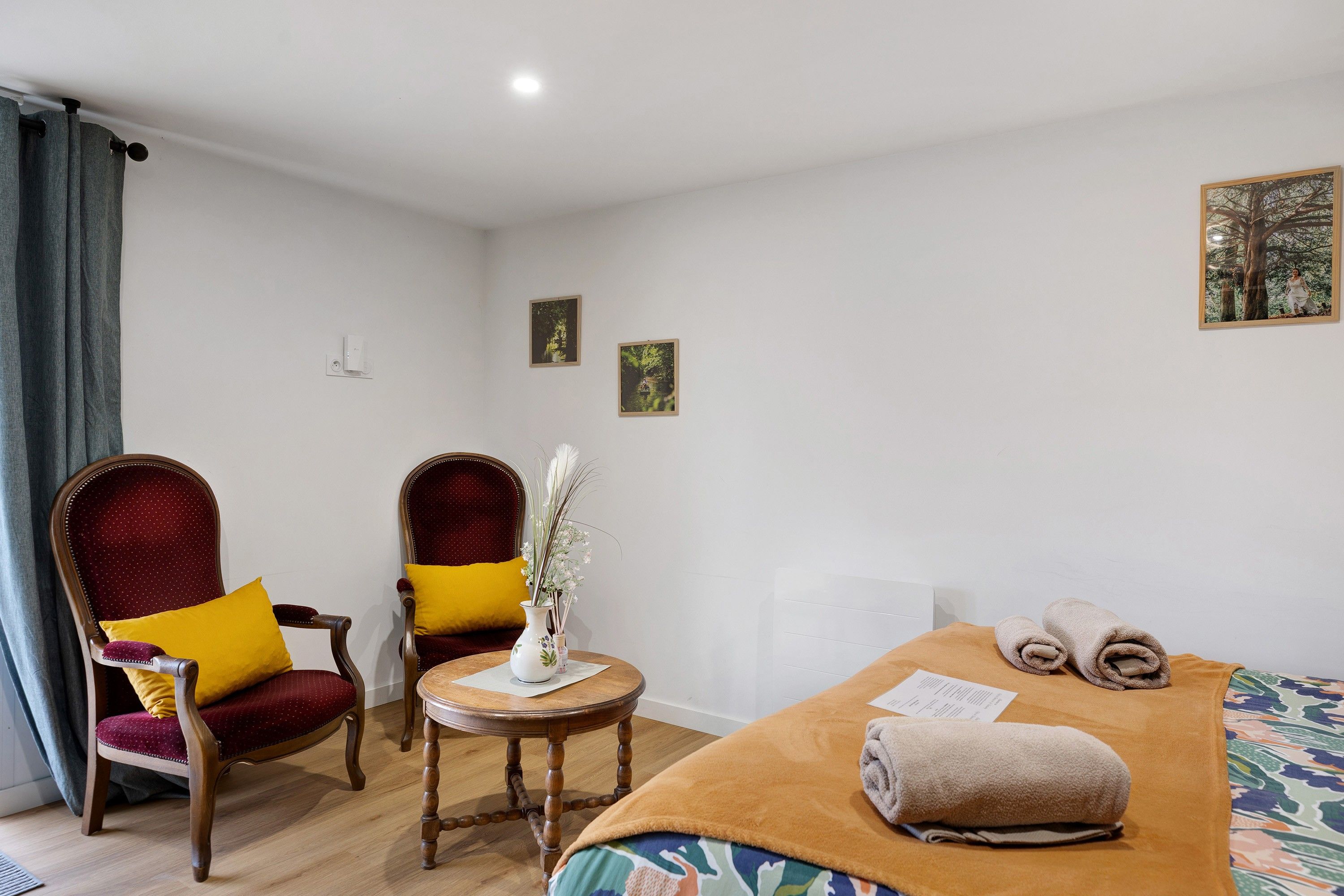 Habitación de hotel con dos sillones, una mesita, una cama con una toalla enrollada y dos cuadros en la pared.