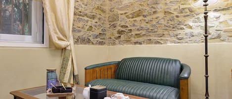 Una habitación con pared de piedra, un sofá de cuero verde, una mesa de madera con tetera y tazas, y una lámpara de pie.