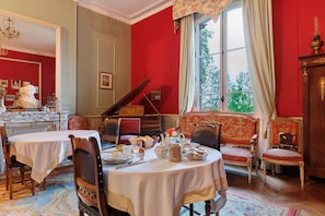 A dining room with a table set for a meal, chairs, a piano, and a window with curtains. - Relai du Château - Canopé PMR Studio (Chemillé-en-Anjou)