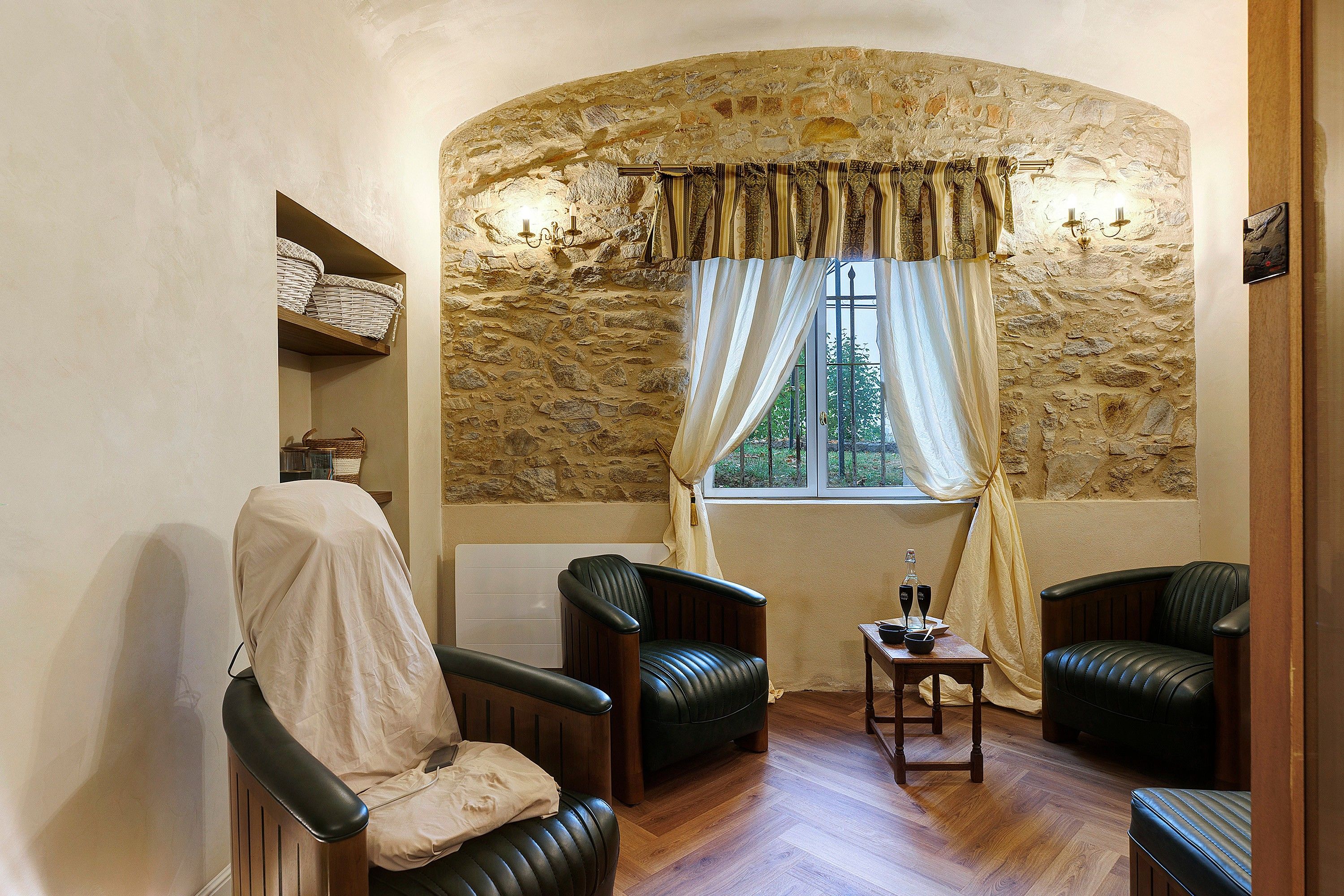 A cozy room with stone walls, a window with curtains, two leather chairs, a small table with a bottle, and a radiator.