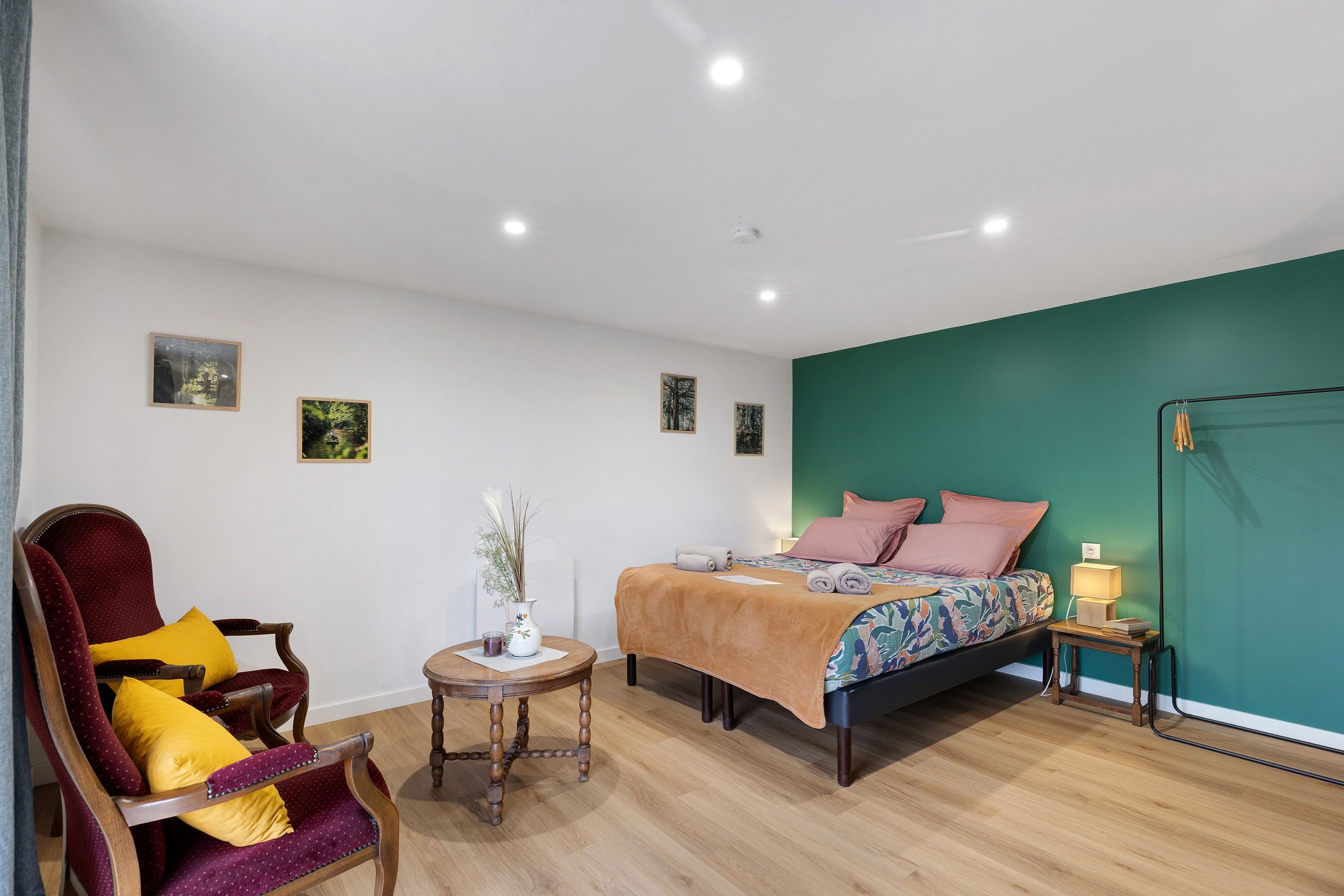 Een slaapkamer met een bed, een stoel, een klein tafeltje met een vaas en een nachtkastje met een lamp.